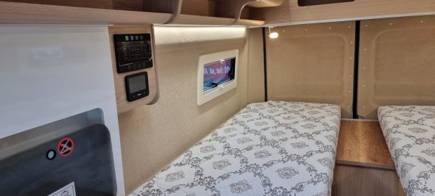 Wnętrze kampera: dwa pojedyncze łóżka z wzorzystymi materacami, małe okno i półka.