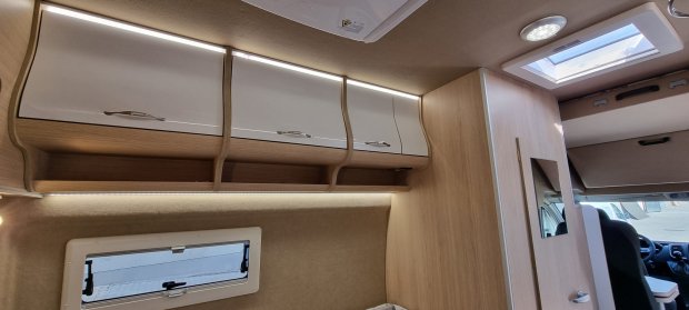 Wnętrze kampera z drewnianymi szafkami, białymi frontami i oknami dachowym oraz bocznym.