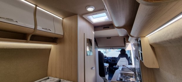 Wnętrze kampera z jasnym drewnem, kuchenką, zlewem, szafkami i przednimi fotelami kierowcy.