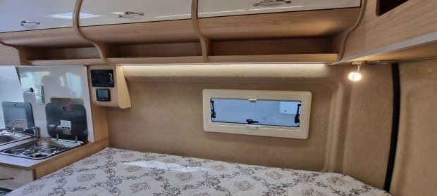 Wnętrze kampera: łóżko przy okienku, półki nad głową i mała kuchnia