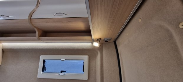 Wnętrze kampera z drewnianą półką, paskiem LED, małą lampką i prostokątnym oknem