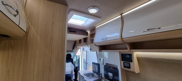 Wnętrze kampera z drewnianymi szafkami, zlewem, świetlikiem i fotelem kierowcy