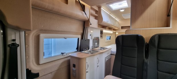 Wnętrze kampera z aneksem kuchennym, zlewem, kuchenką, szafkami i łóżkiem