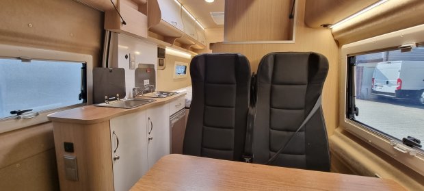 Wnętrze kampera: kuchnia ze zlewem i szafkami oraz dwa czarne fotele przy stole