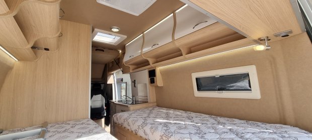 Wnętrze kampera z dwiema kojami, drewnianymi szafkami i małym oknem