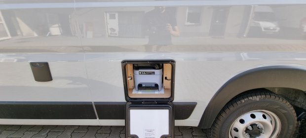 Boczna klapa kampera otwarta, widoczna kasetowa toaleta w schowku nad kołem.