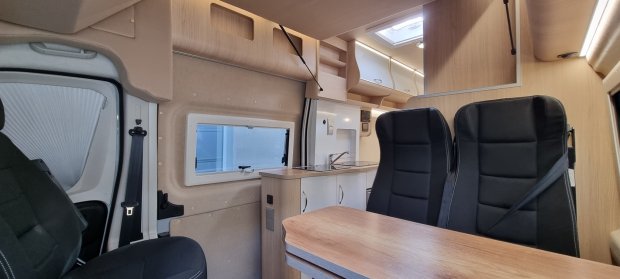 Wnętrze kampera z drewnianą zabudową, stołem i dwiema czarnymi fotelami.