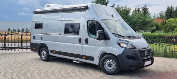 Szary kamper Fiat LUB-CAMP zaparkowany przy ozdobnej metalowej bramie.