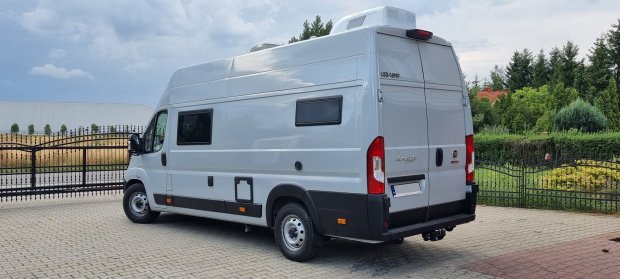 Biały kamper van zaparkowany na podjeździe przy metalowej bramie