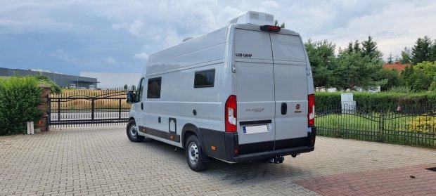 Biały kamper typu bus zaparkowany przy bramie na brukowanym podjeździe.