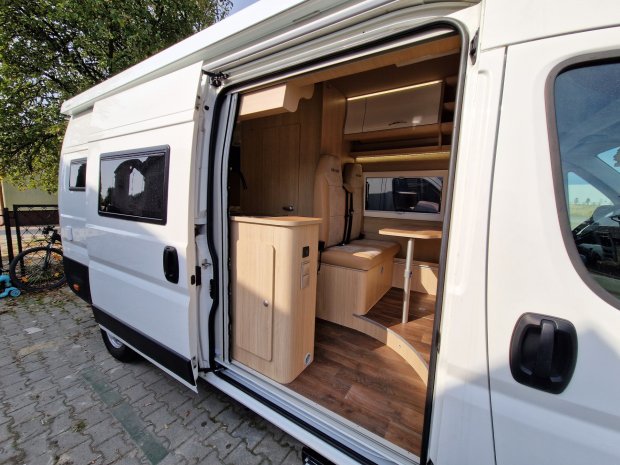 Otwarty biały kamper, widoczne drewniane wnętrze z siedziskami i stolikiem
