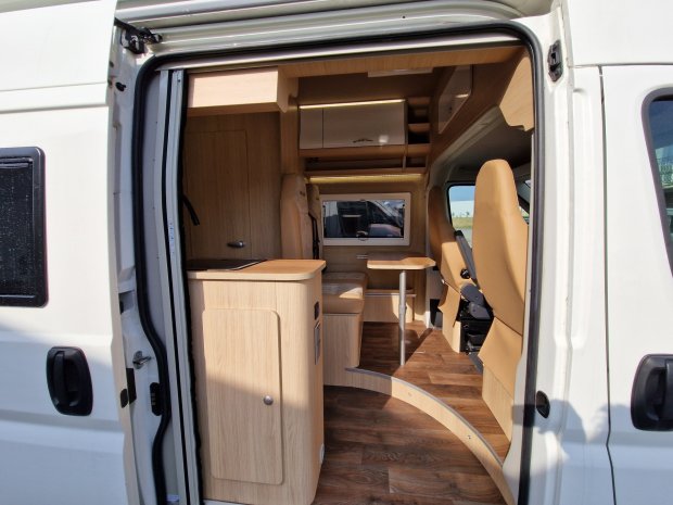 Wnętrze kampera z drewnianymi szafkami, beżowymi fotelami i małym stołem.