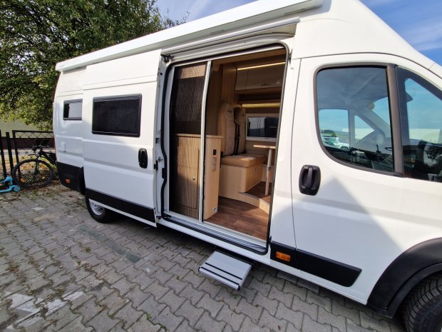 Biały kamper z otwartymi drzwiami bocznymi i widocznym drewnianym wnętrzem.