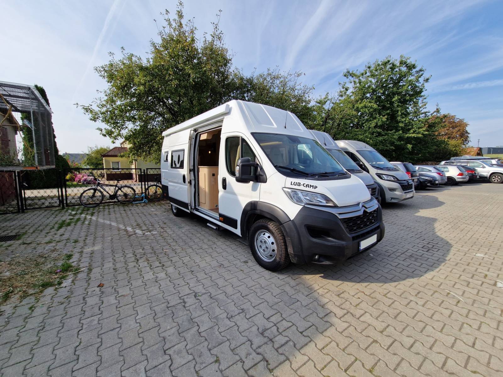 Biały kamper z otwartymi drzwiami zaparkowany na placu parkingowym.