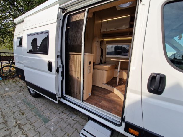 Biały kamper z otwartymi drzwiami, widoczne wnętrze z siedzeniem i stolikiem