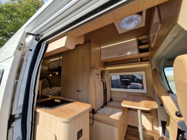 Wnętrze kampera z jasną drewnianą zabudową, aneksem kuchennym, siedzeniami i stołem.
