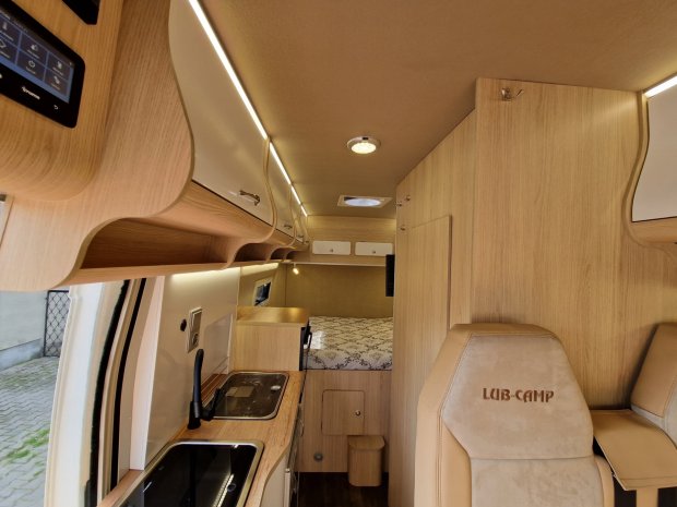 Jasne wnętrze kampera z drewnianą zabudową, aneksem kuchennym, łóżkiem i fotelem LUB-CAMP.