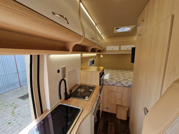 Wnętrze kampera: kuchnia z zlewem, płytą i drewnianymi szafkami oraz łóżko.