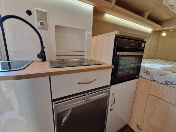 Wnętrze przyczepy: mała kuchnia z czarną baterią, zlewem, piekarnikiem i łóżkiem.