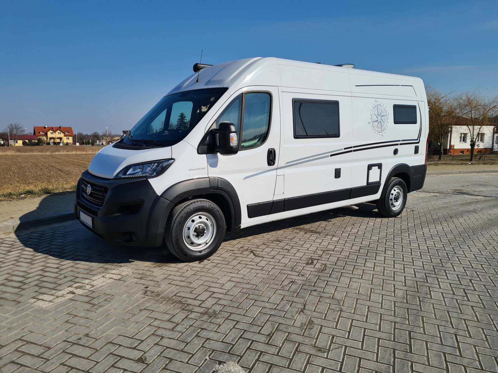 Biały kamper van zaparkowany na kostce brukowej przy polu i domach.