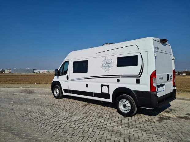 Biały kamper z kompasem na boku zaparkowany na brukowanym placu przy bezchmurnym niebie