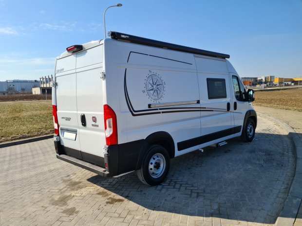 Biały kamper Fiat z kompasową grafiką na boku, zaparkowany na brukowanym placu.