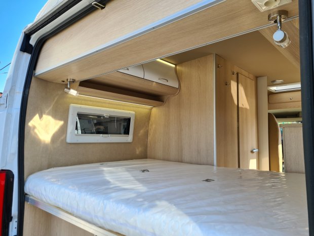 Wnętrze kampera z drewnianymi szafkami, łóżkiem i małym oknem.