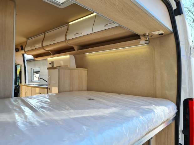 Wnętrze kampera z łóżkiem, drewnianymi szafkami i małą kuchnią