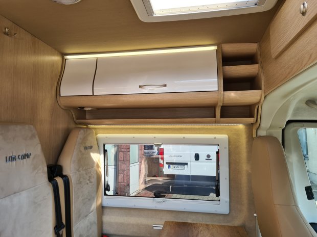 Wnętrze kampera z drewnianymi szafkami, białą półką i oknem nad siedziskiem.