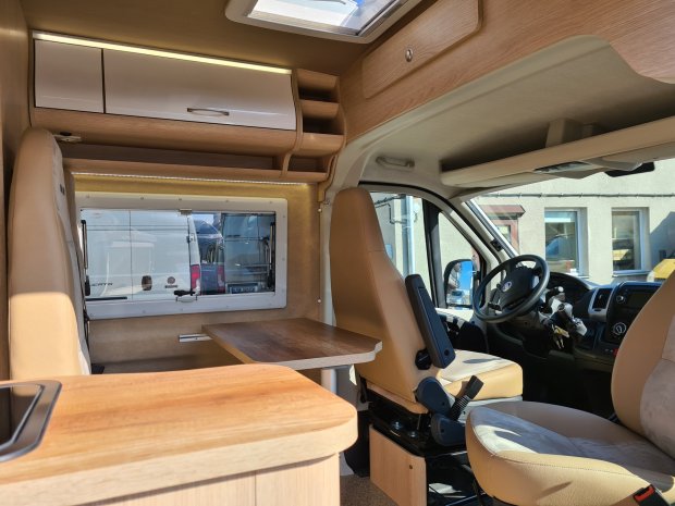 Wnętrze kampera: przednie fotele w beżu, stolik i drewniane szafki.