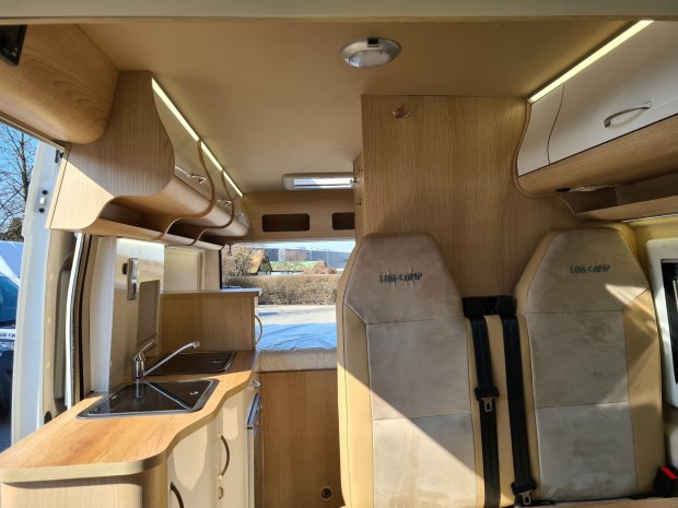 Jasne, drewniane wnętrze kampera z kuchenką, szafkami i dwoma fotelami.