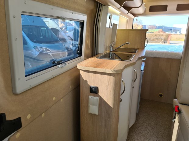 Wnętrze kampera z drewnianą kuchnią, podwójnym zlewem, oknem i łóżkiem z tyłu.