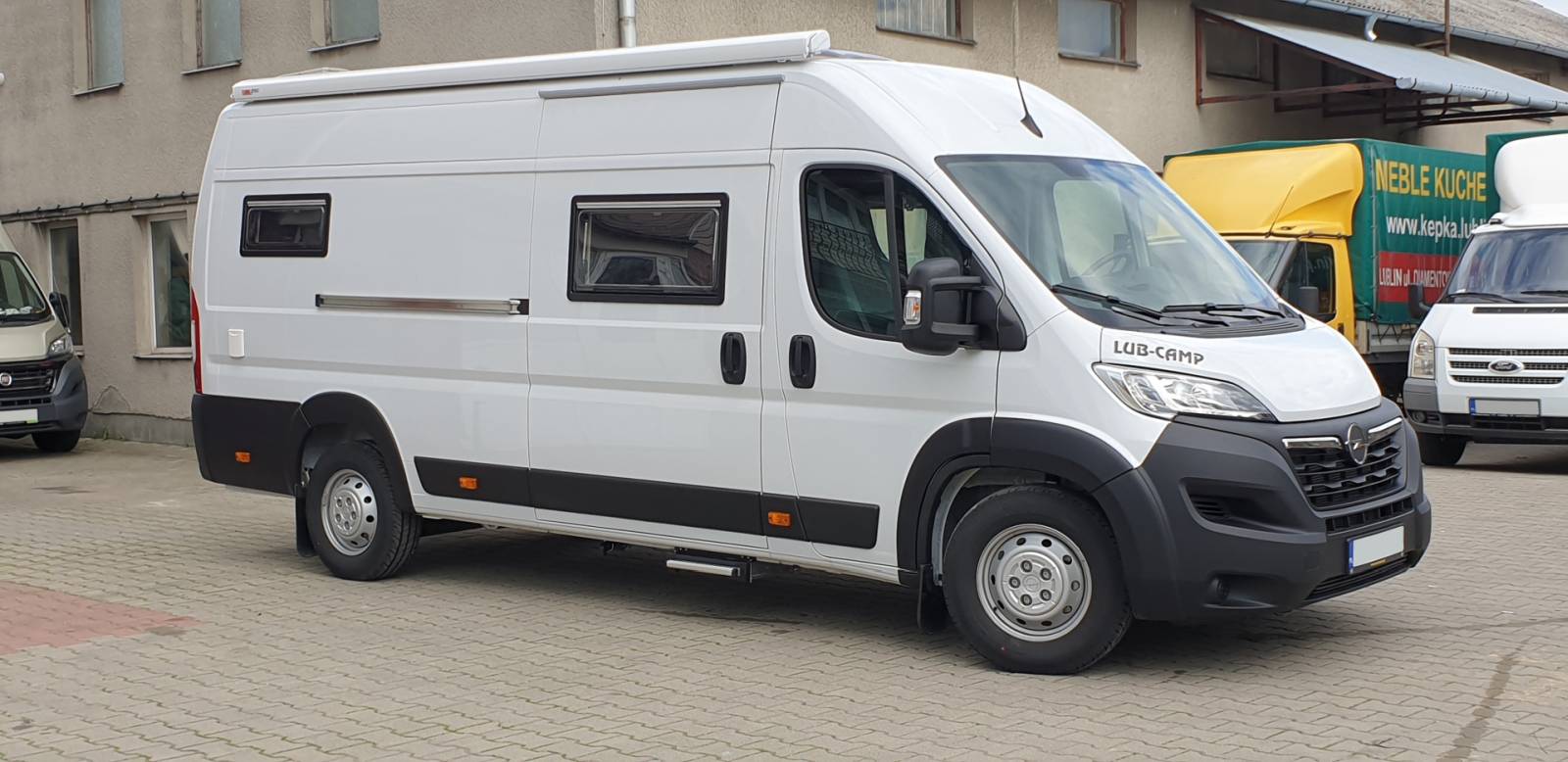 Biały camper-van zaparkowany przy budynku, widok z prawego boku