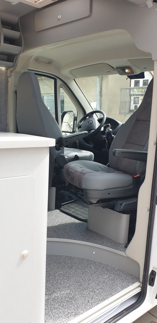 Wnętrze kampera z obróconymi fotelami kierowcy i pasażera, szarymi siedziskami i szafką.