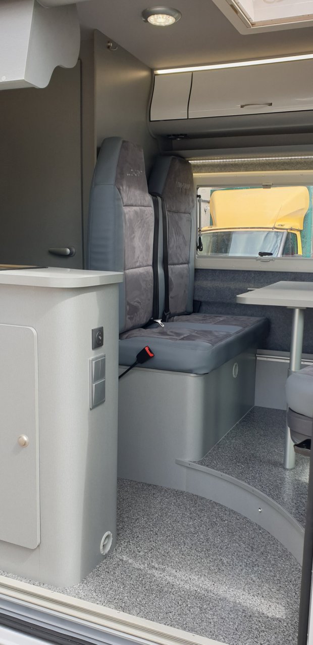 Wnętrze kampera: szare tapicerowane siedzenia, stolik i zabudowa kuchenna.