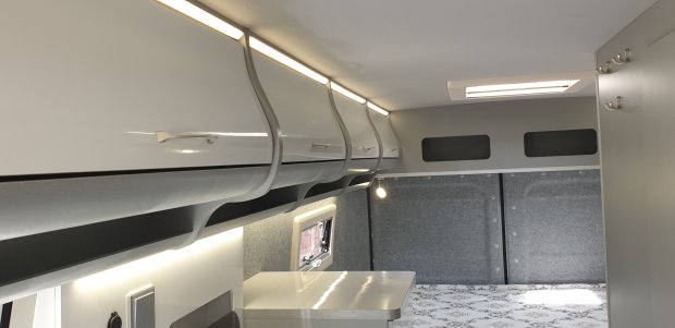 Wnętrze kampera z białymi szafkami nad głową, oświetleniem LED i łóżkiem.
