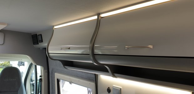 Górne jasnoszare szafki i półka z oświetleniem we wnętrzu kampera