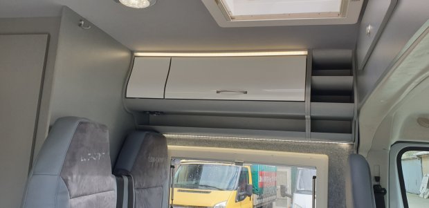 Górna szafka i półki nad siedzeniami kampera, przez okno widać żółtą ciężarówkę.