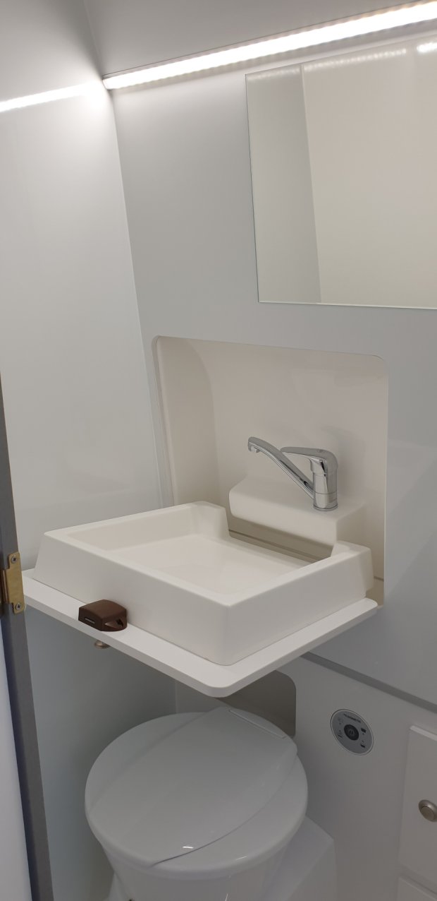 Mała biała umywalka z chromowaną baterią nad zamkniętą toaletą i lustrzaną półką.