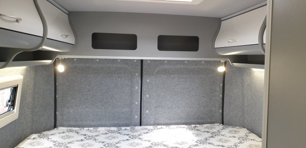 Wnętrze kampera: łóżko z wzorzystą pościelą, szare panele, półki i lampki