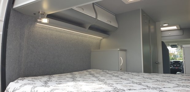 Wnętrze kampera: łóżko z wzorzystą pościelą, szare szafki, półki i oświetlenie LED.