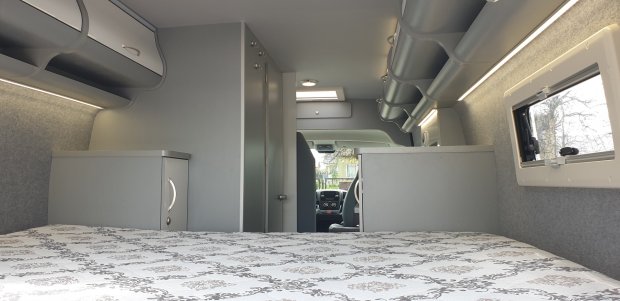 Wnętrze kampera: łóżko z wzorzystą pościelą, szare szafki i okno