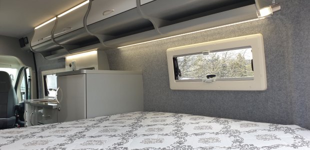 Wnętrze kampera: łóżko z wzorzystą pościelą, szafki, okienko i mini kuchnia