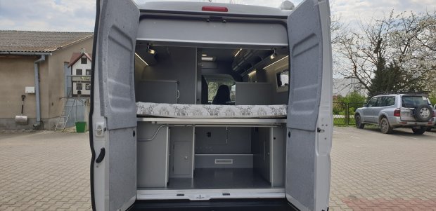 Tył kampera z otwartymi drzwiami, łóżkiem i schowkami