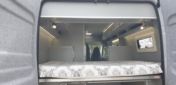 Wnętrze kampera z rozkładanym łóżkiem, górnymi schowkami i małym okienkiem.