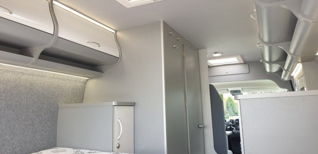 Wnętrze kampera z jasnoszarymi szafkami, półkami nad łóżkiem i widokiem na kabinę.