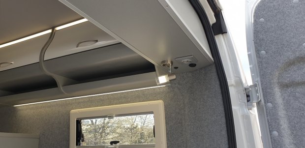 Wnętrze kampera z półkami sufitowymi, taśmami LED, lampką i oknem.