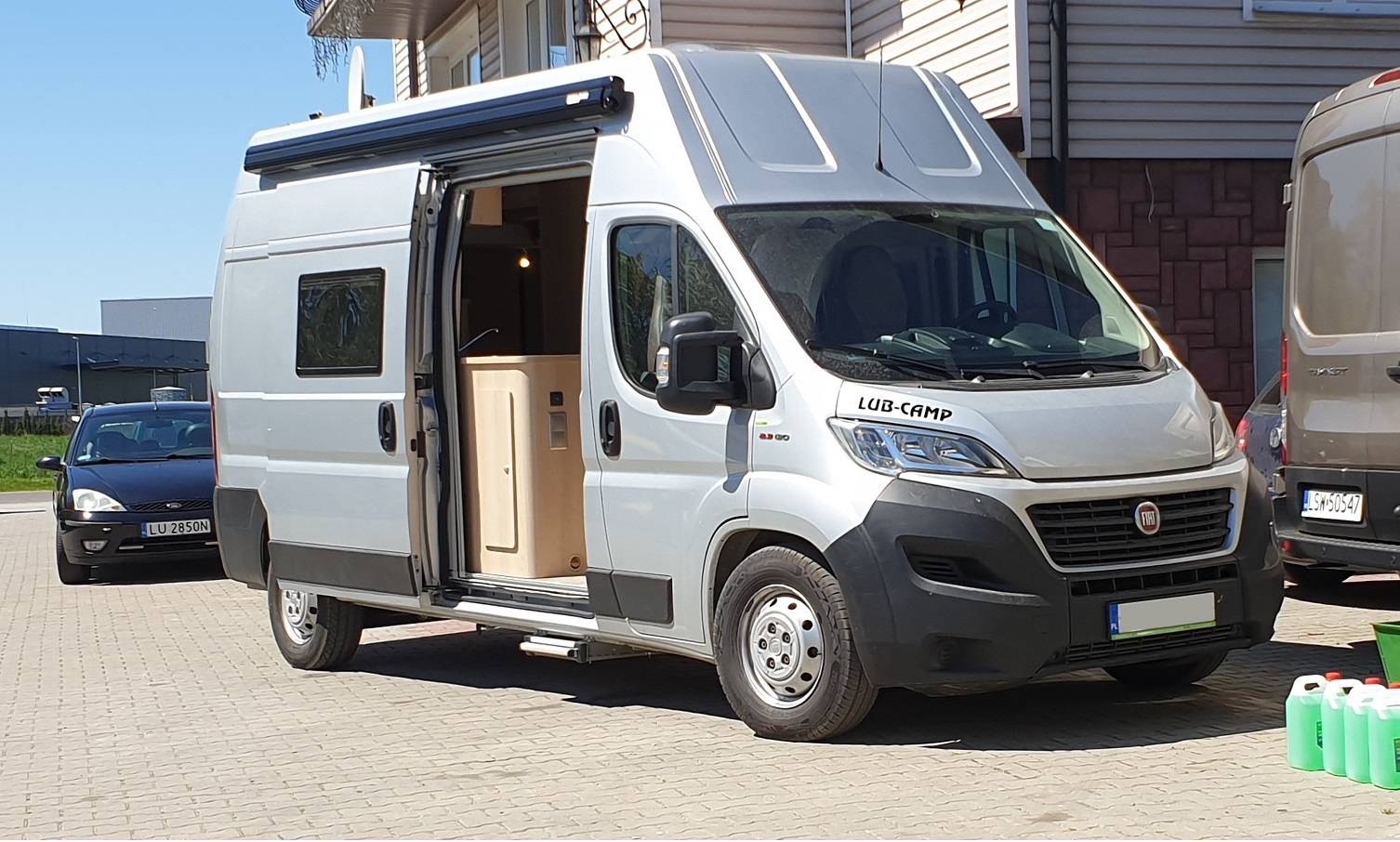Biały kamper Fiat z otwartymi drzwiami bocznymi zaparkowany przed budynkiem