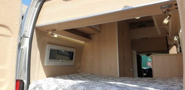 Wnętrze kampera: łóżko z jasną pościelą, drewniane szafki i lampki.