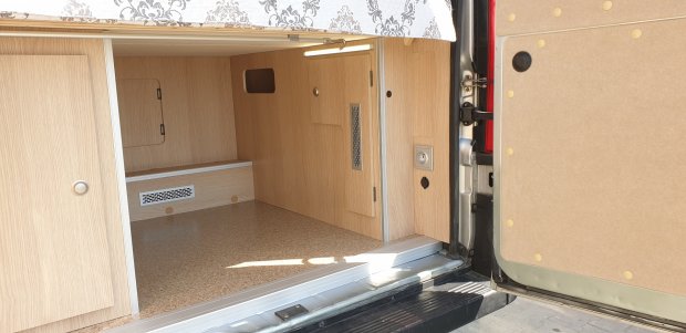 Otwarty schowek kampera z jasnymi drewnianymi panelami, oświetleniem i wykładziną.